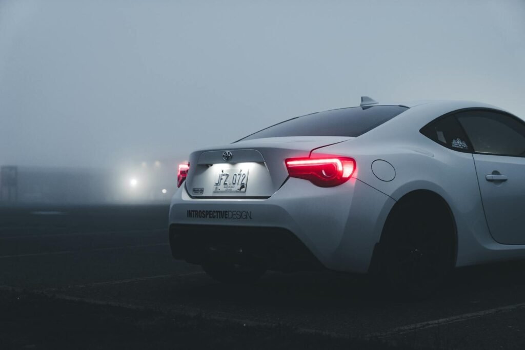White sports car with illuminated taillights in a foggy urban parking lot at dusk.