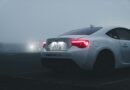 White sports car with illuminated taillights in a foggy urban parking lot at dusk.
