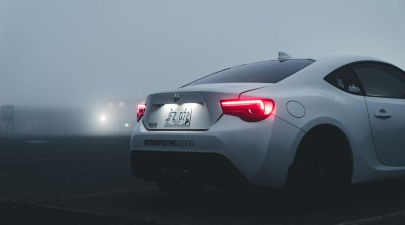 White sports car with illuminated taillights in a foggy urban parking lot at dusk.